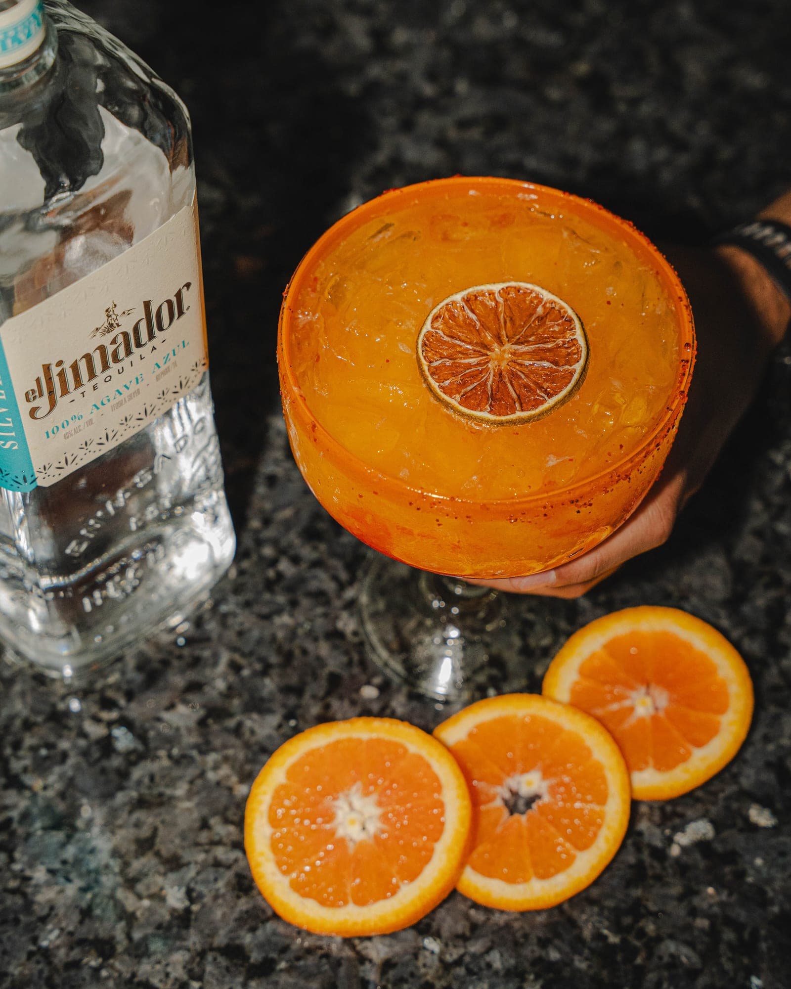 An orange margarita with a chile-salt rim and a dried orange wheel, next to a bottle of el Jimador tequila and fresh orange slices on dark granite